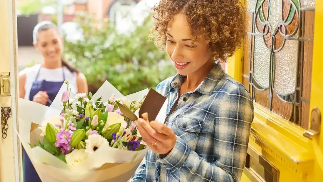 Woman receiving flower delivery, smiling and reading card at her door.