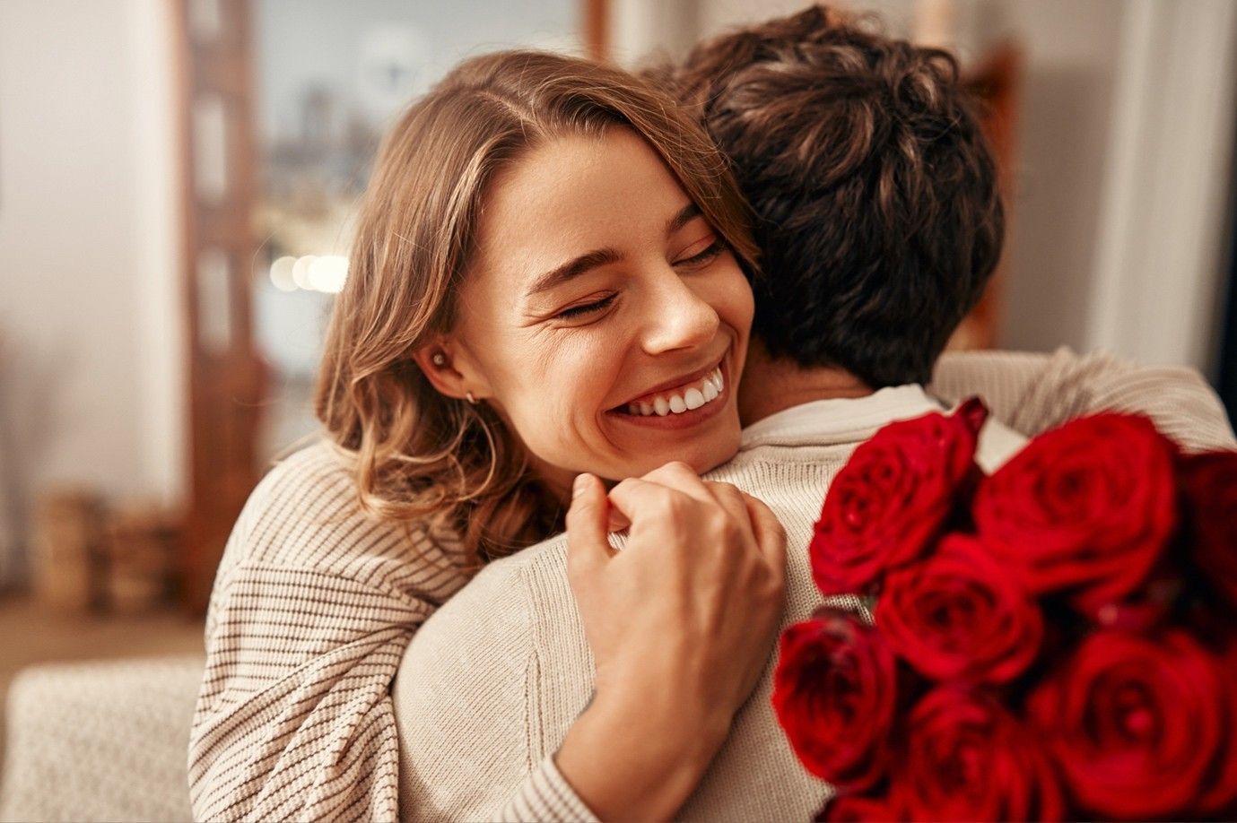 a woman is hugging a man with a bouquet of red roses behind his back .