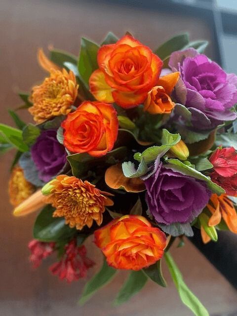 Bouquet of orange roses, purple kale flowers, and green leaves against a blurry background.