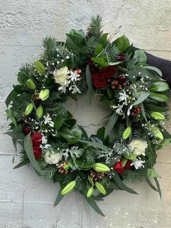 Christmas wreath with red roses, white flowers, and green foliage against a white brick wall.