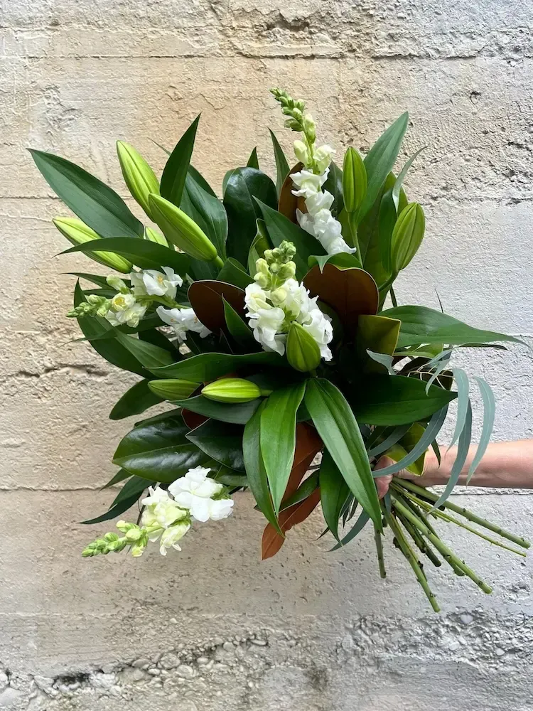 Bouquet of white snapdragons, lilies, and green leaves held against a textured wall.