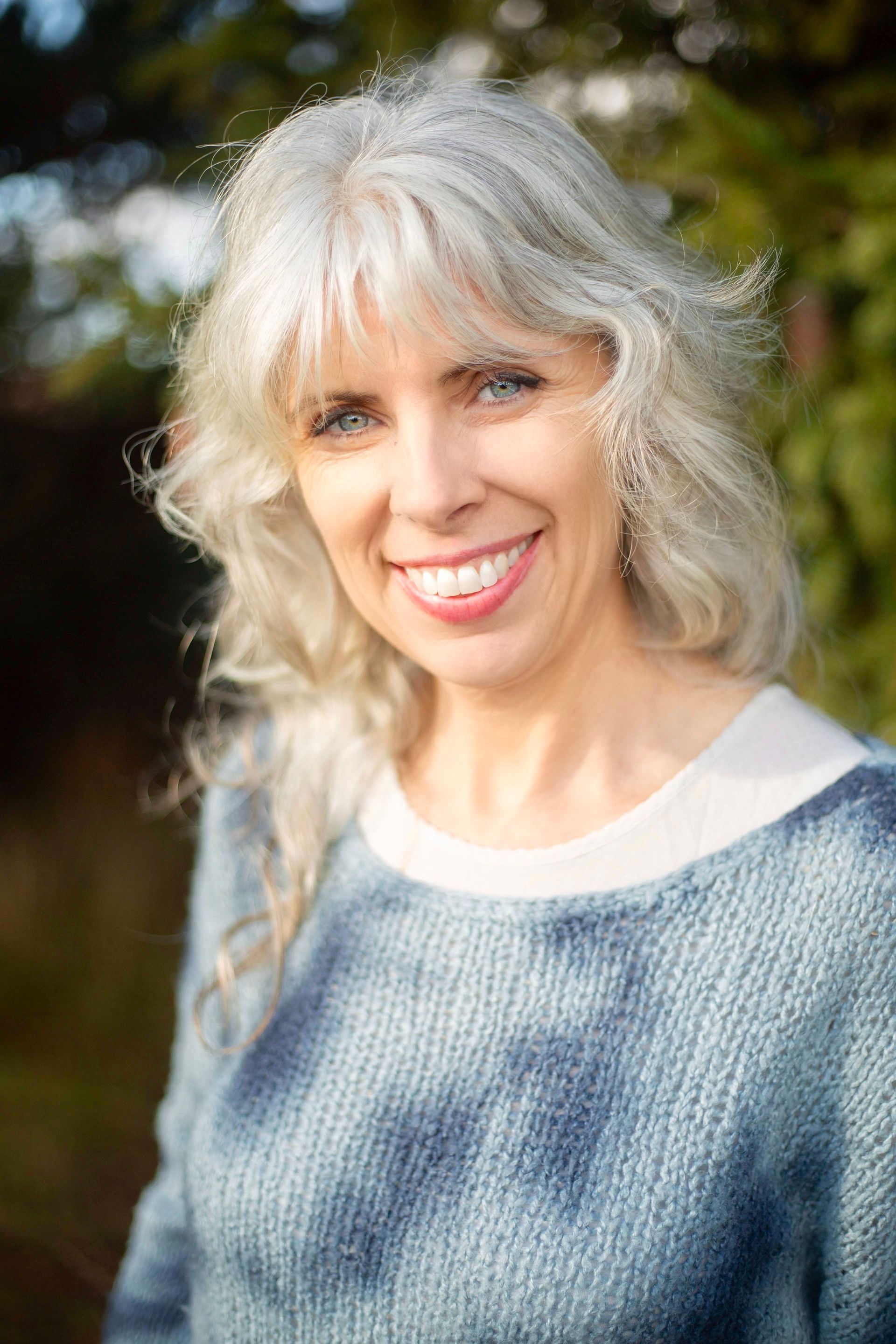 Woman with gray hair smiles, wearing a blue sweater, against a blurred green background.