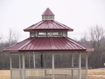 A maroon-roofed gazebo with white posts and railings stands in a field