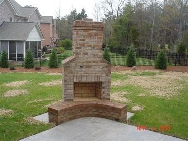 Brick outdoor fireplace with curved seat on a concrete patio, grassy yard, and houses in the background