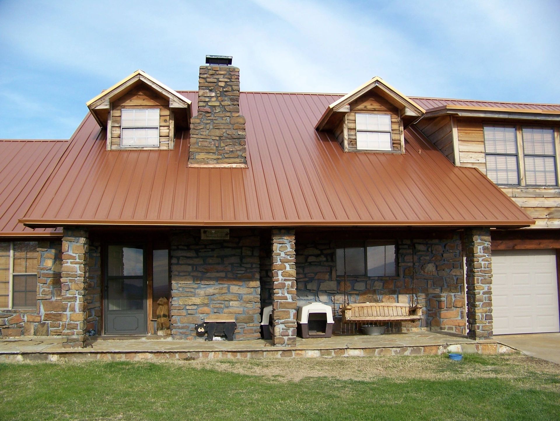 Brown-roofed stone house with dormers, chimney, and porch, against a blue sky