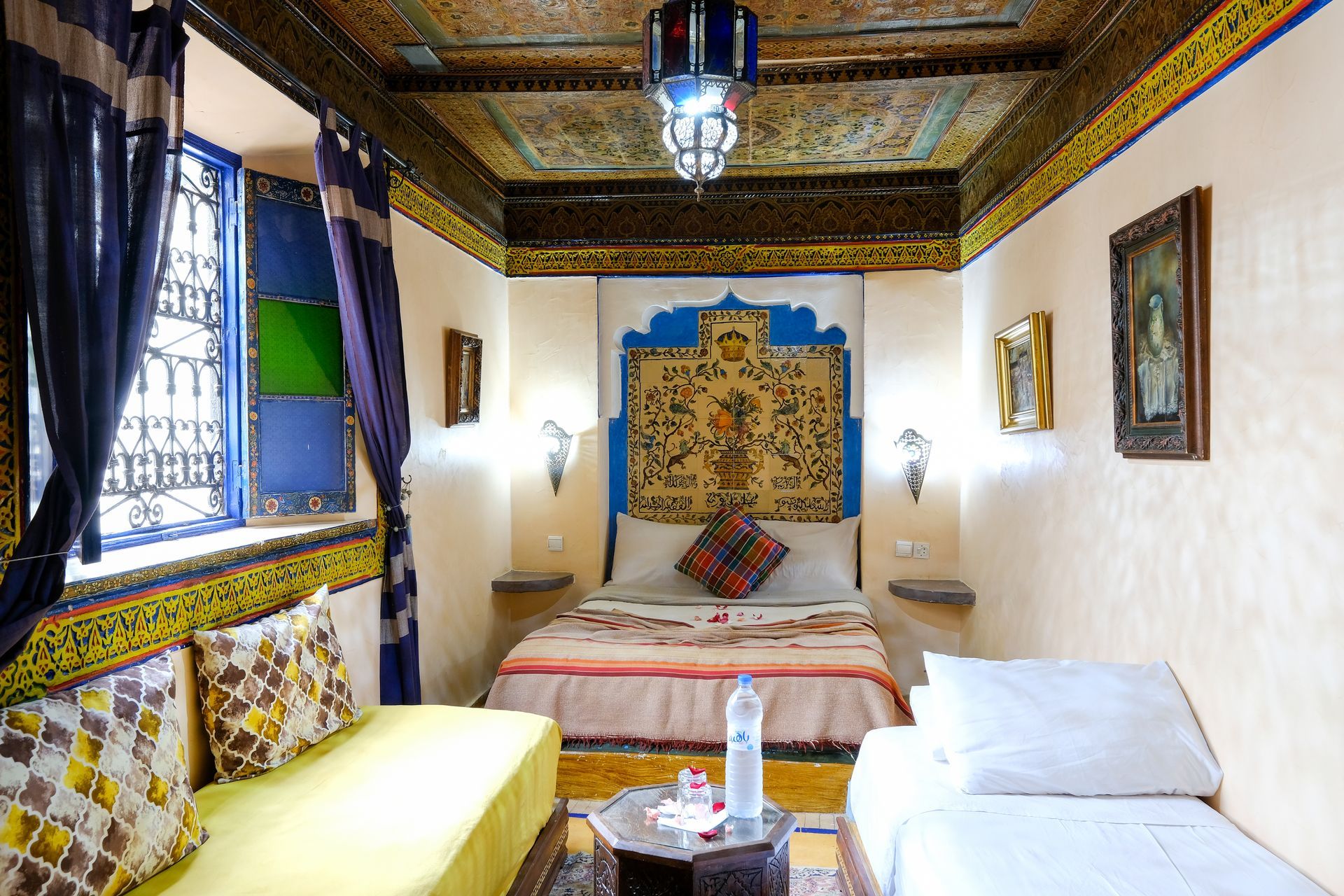 Moroccan-style bedroom with ornate ceiling, colorful details, window with blue shutters, and seating area.