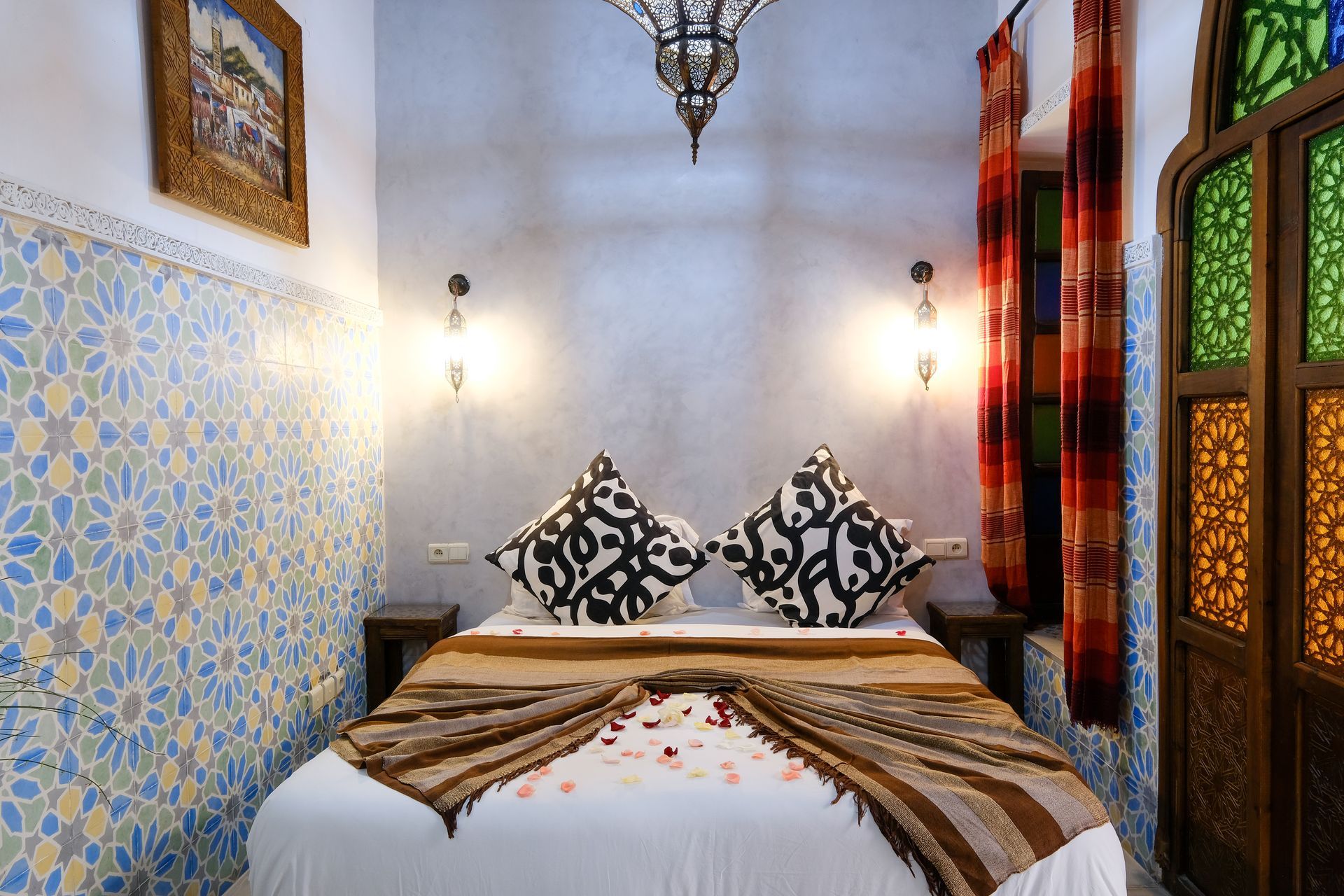 Bedroom with a bed, patterned pillows, blue tiled wall, stained glass window, and ornate pendant light.