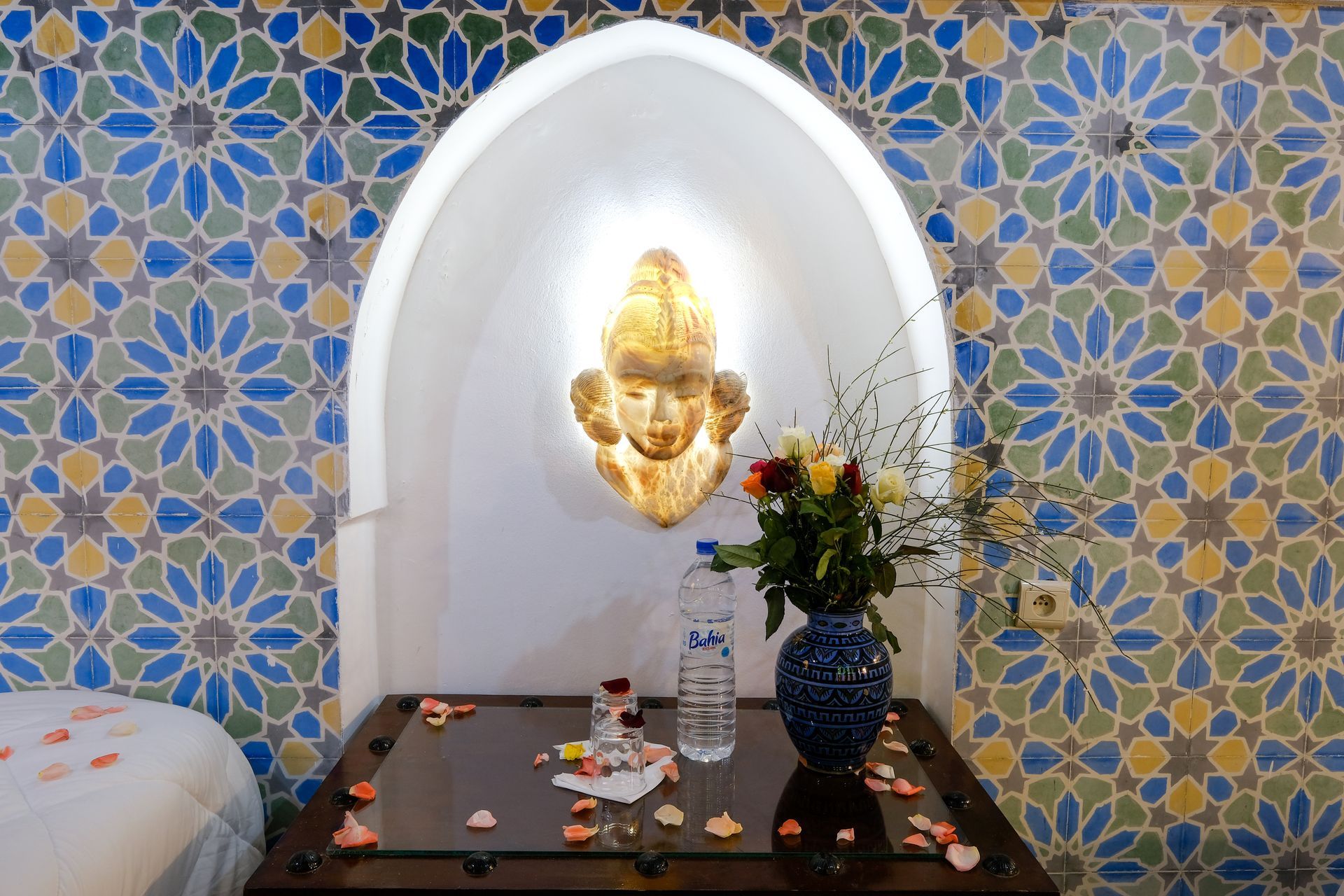Room with mosaic tile wall, arched alcove, and table with flowers and water bottles.