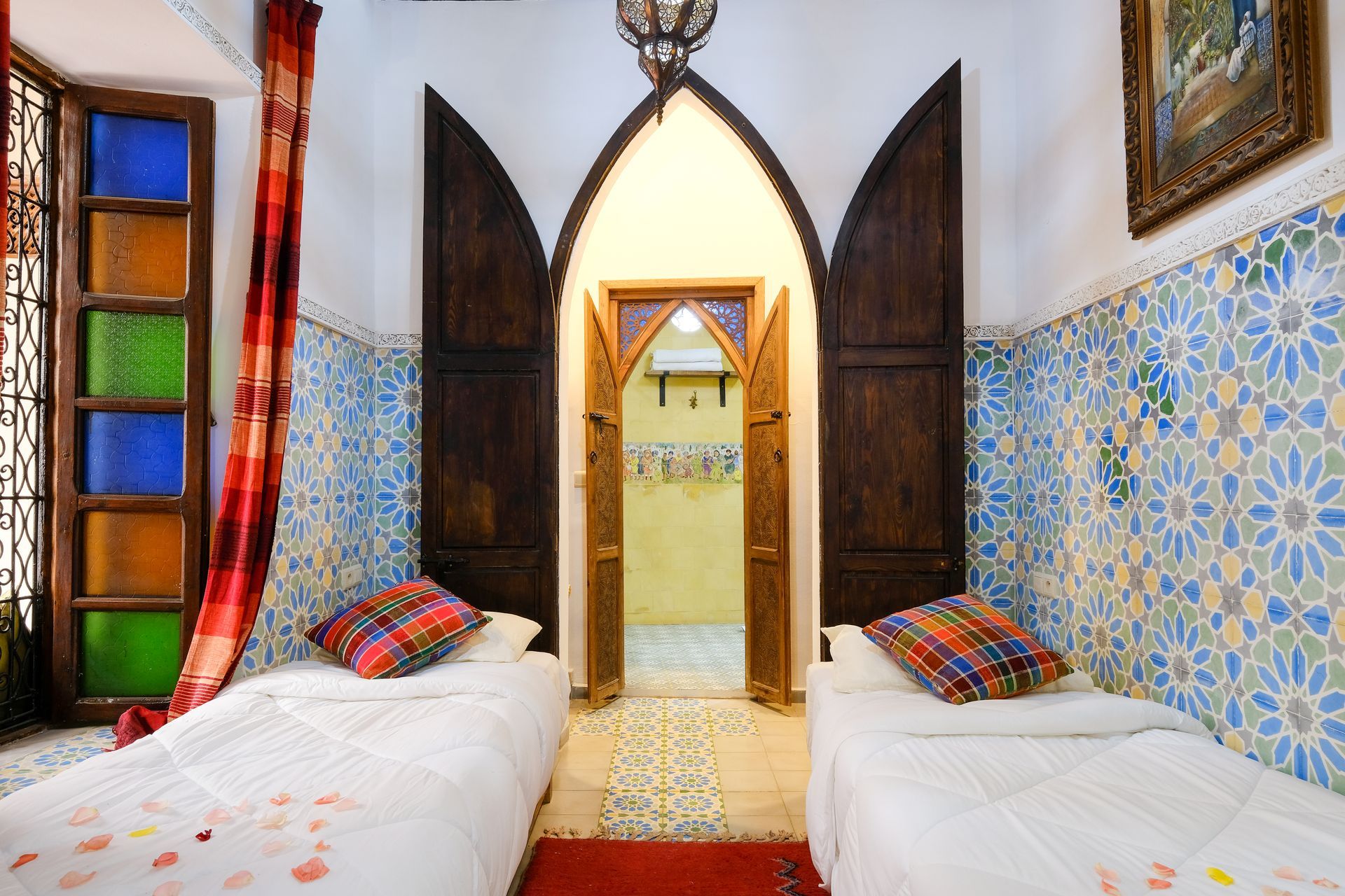 Twin beds in a Moroccan-style room, patterned blue tile, stained glass window, ornate doorway, red accents.