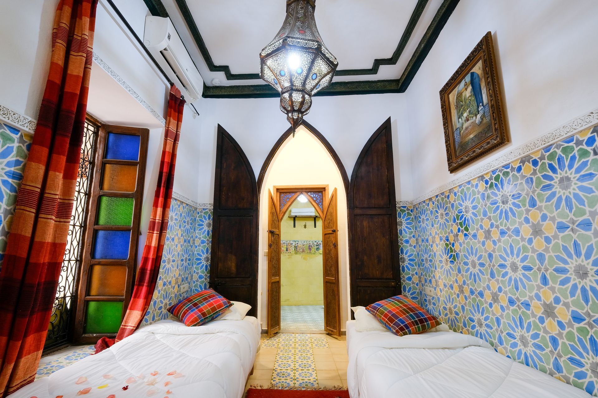 Twin beds in a Moroccan-style room, colorful stained-glass window, floral tile wall, lantern chandelier.