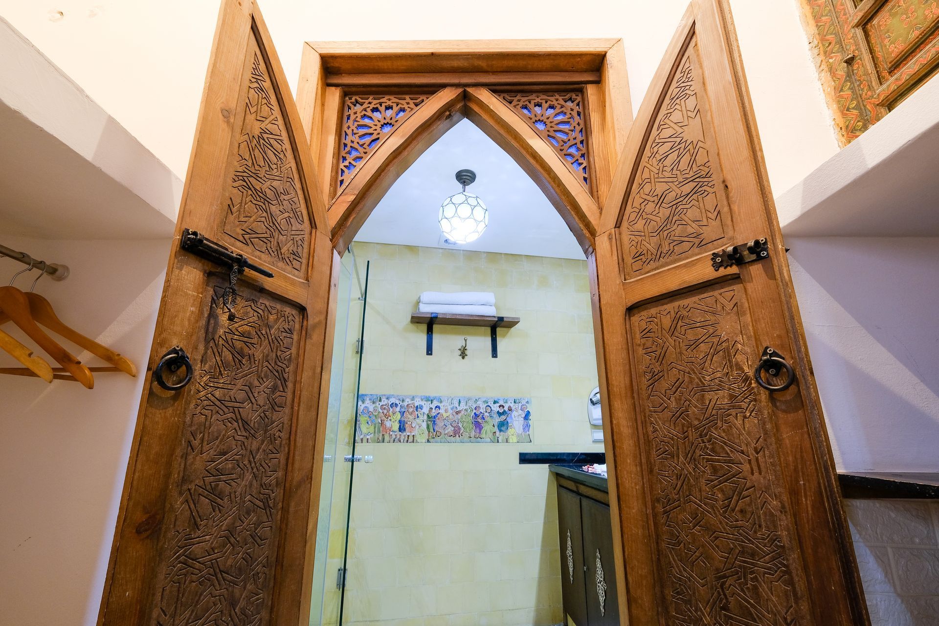 Wooden carved doors open to a brightly lit room; a light fixture hangs in the arched doorway.