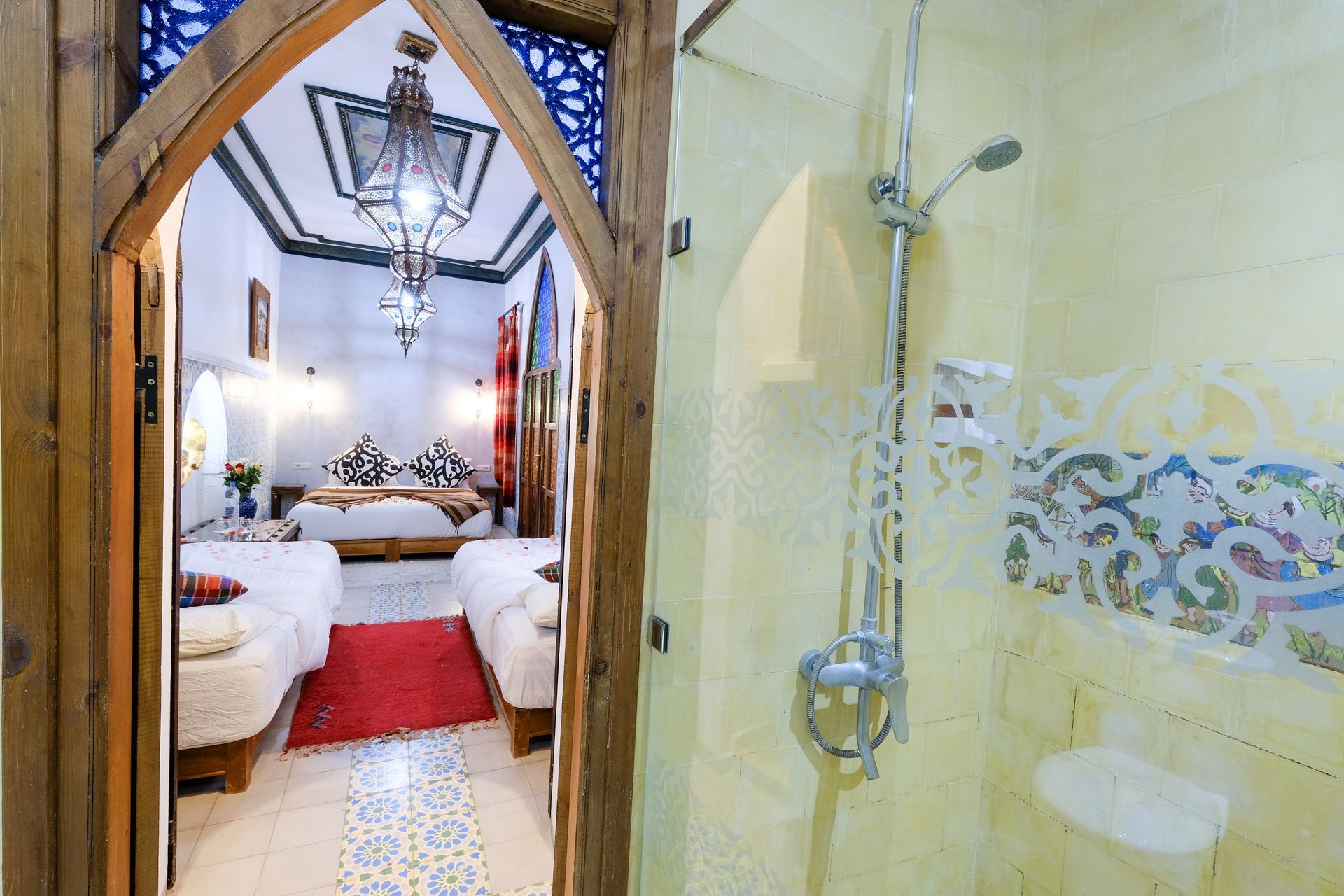 View through an arched doorway into a Moroccan style bedroom. A shower with glass doors is in the foreground.