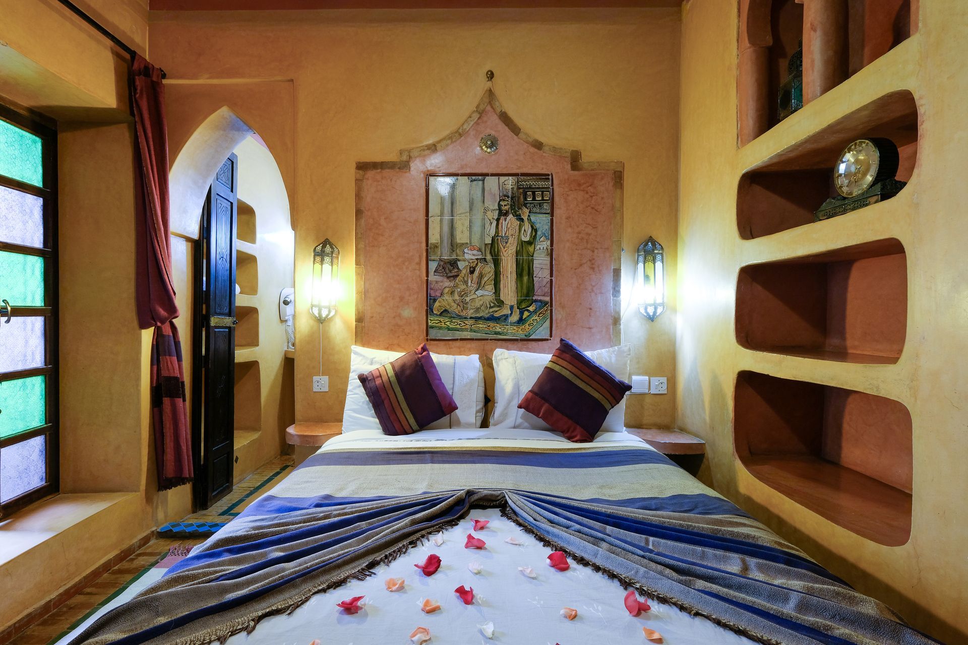 Bedroom with arched doorways, colorful window, decorative bedspread, and built-in shelves.