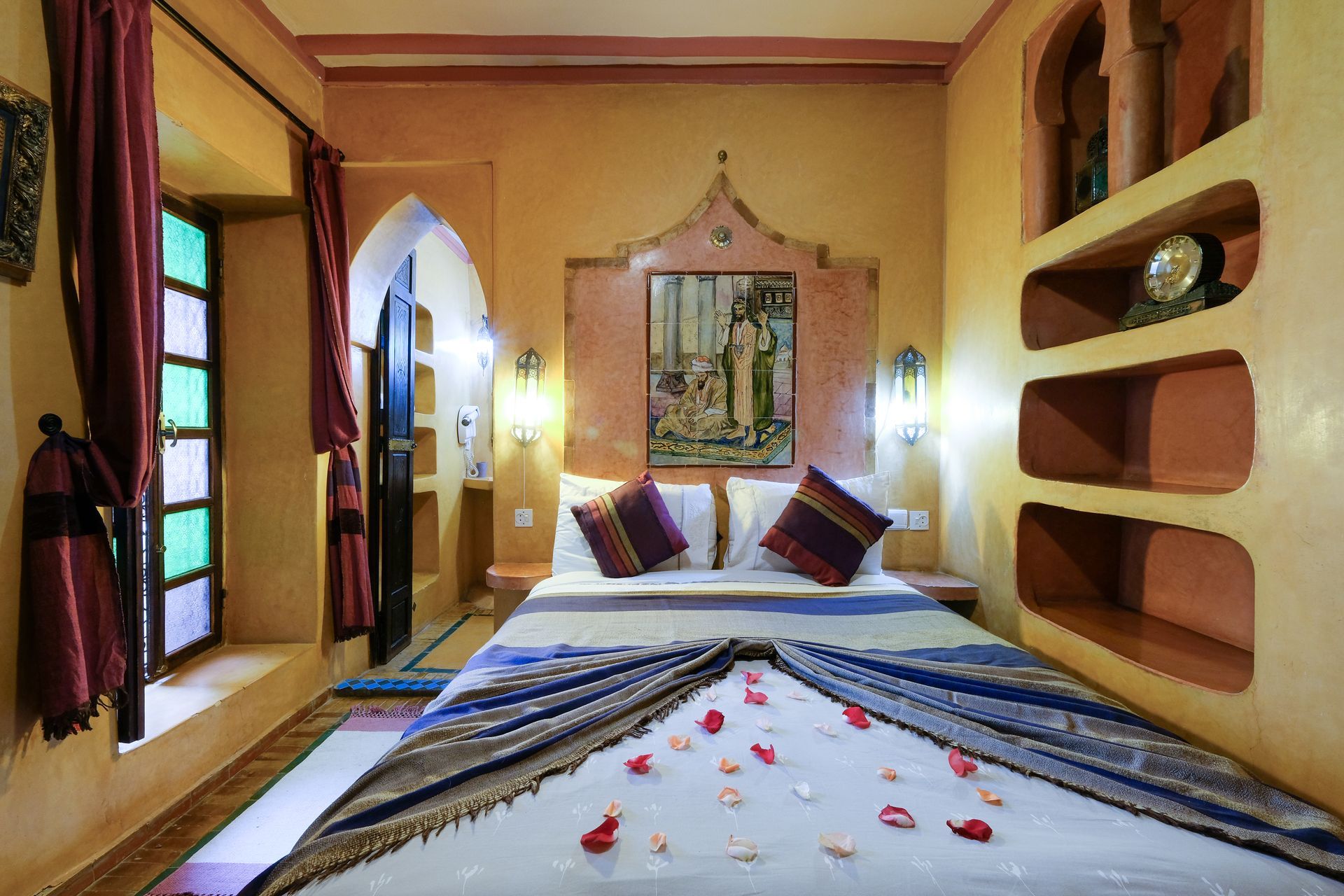 Bedroom in Moroccan style, peach walls, bed with pillows, rose petals, archways, and window with colorful glass.