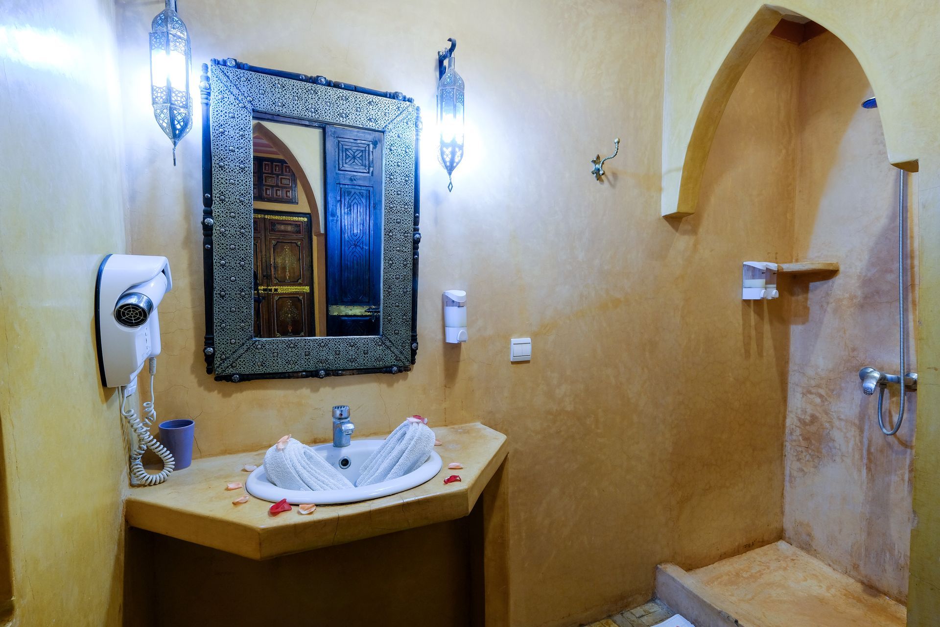 Bathroom with ornate mirror, sink, shower, and arched doorway, in shades of yellow and brown.