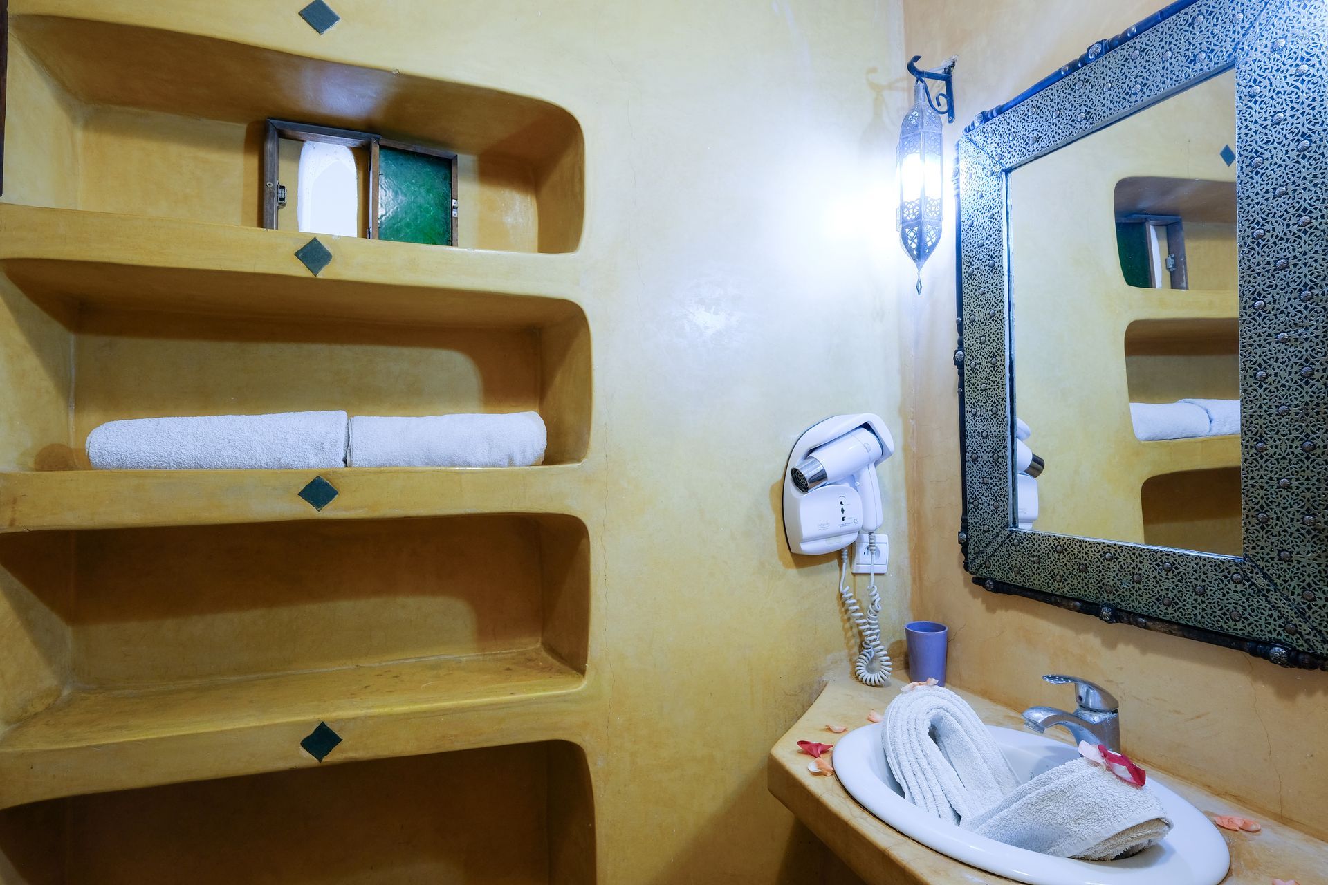 Bathroom with yellow walls, built-in shelves holding towels, sink, mirror with decorative frame.