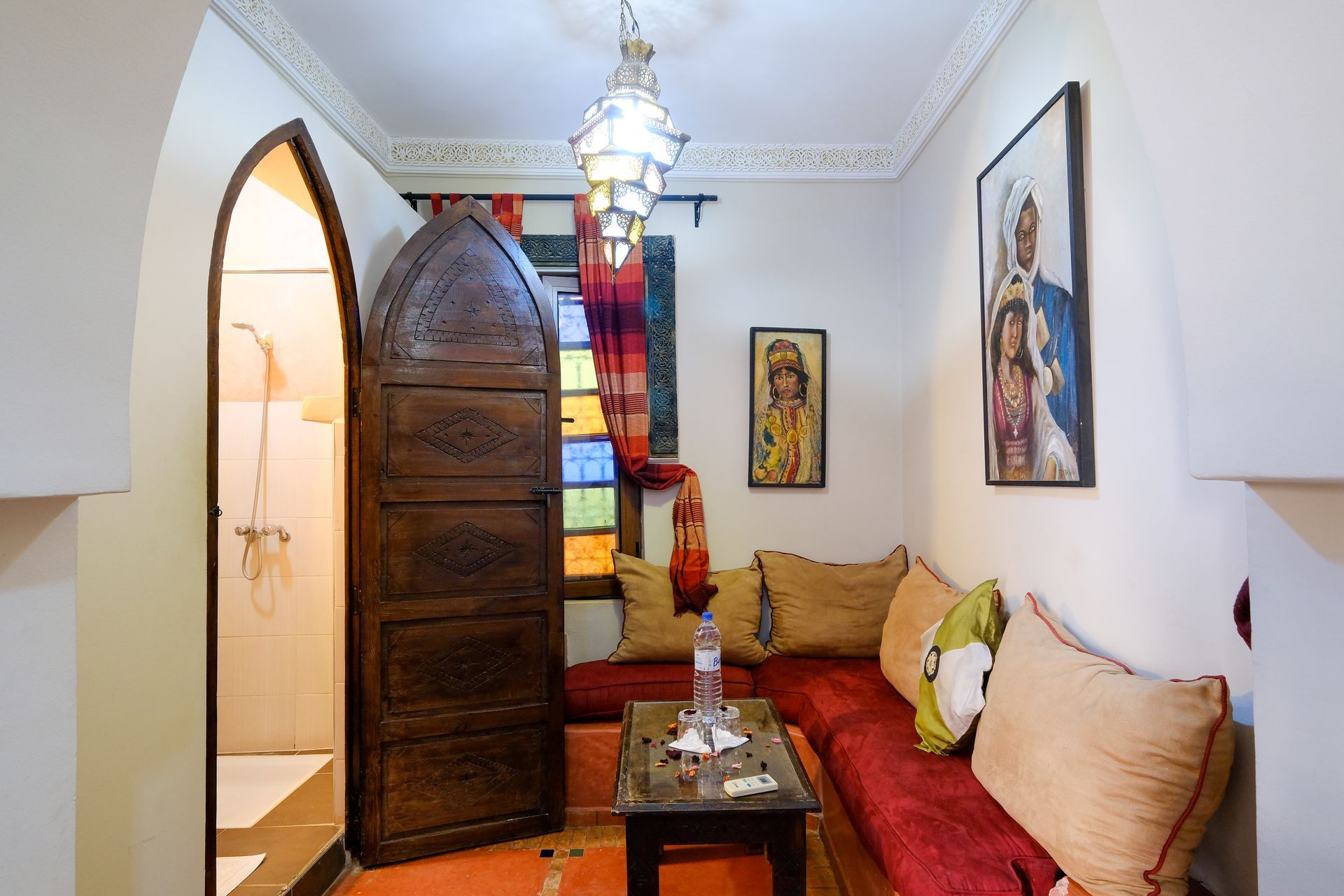 Cozy Moroccan-style room: arched doorway, wooden door, red sofa with pillows, small table, colorful window, framed art, and hanging lantern.