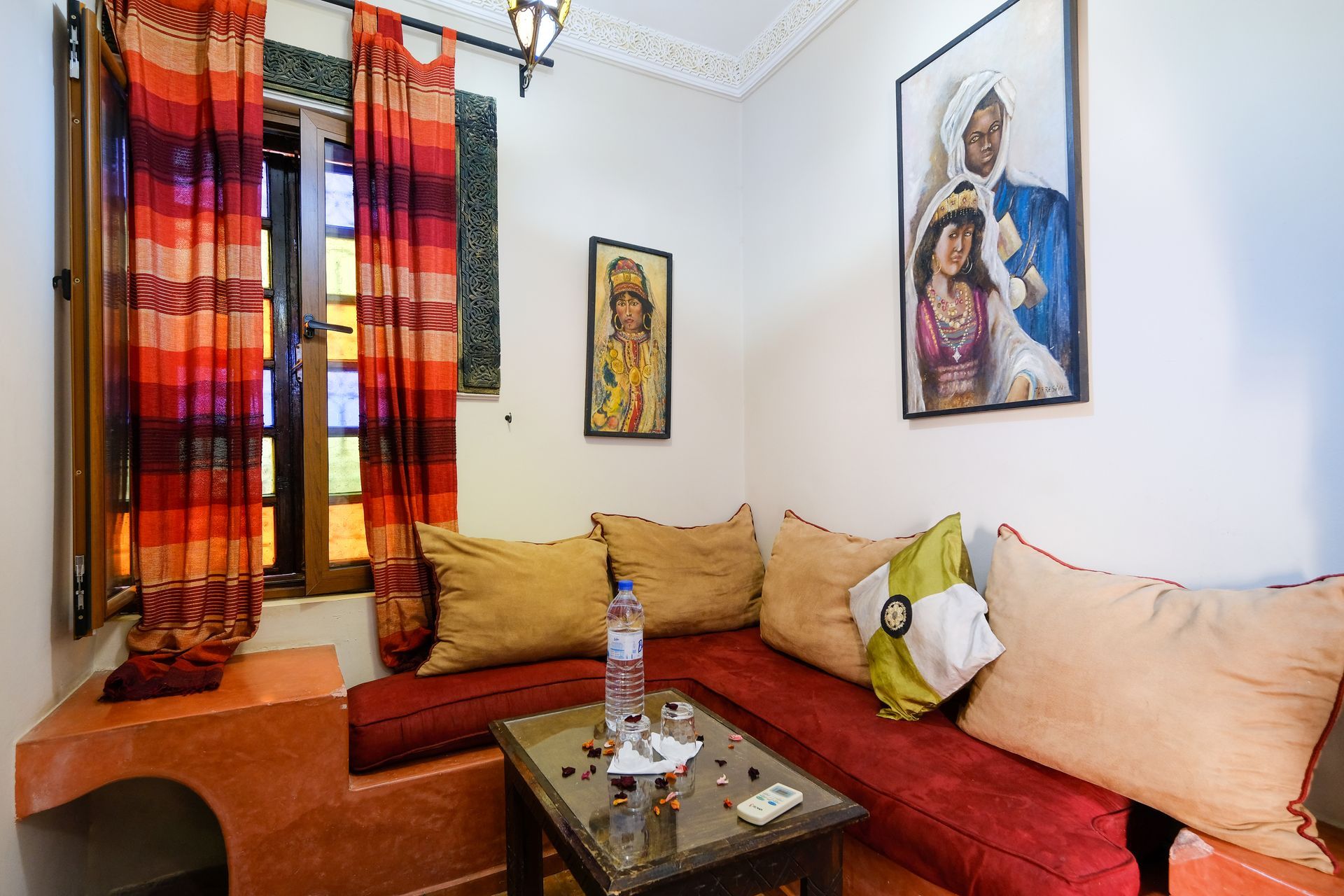 Cozy living area with red built-in seating, cushions, small table, and stained glass window. Paintings on the wall.