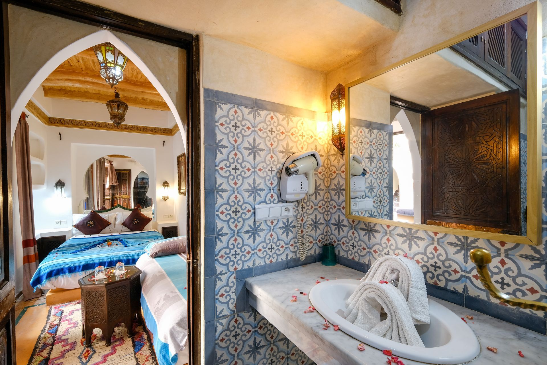 Bathroom with patterned blue tiles, sink, and ornate mirror. Doorway to bedroom with bed.