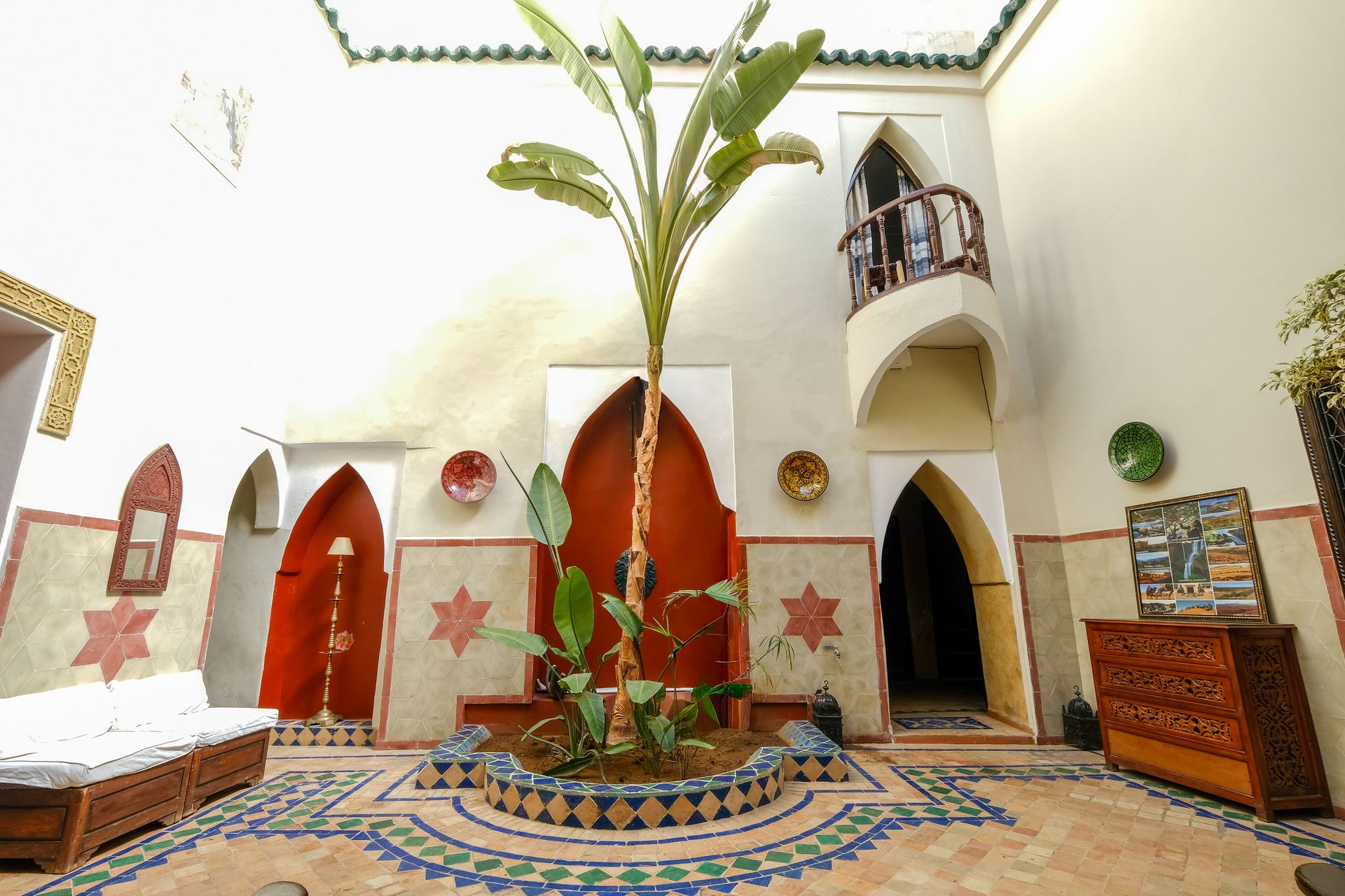 Courtyard with tiled floor, arched doorways, a plant in the center, and a balcony above. White and red walls.