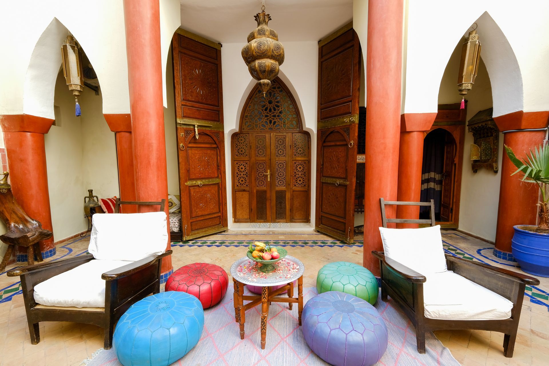 Courtyard with wooden doors, red pillars, white chairs, colorful poufs, and a hanging lantern.