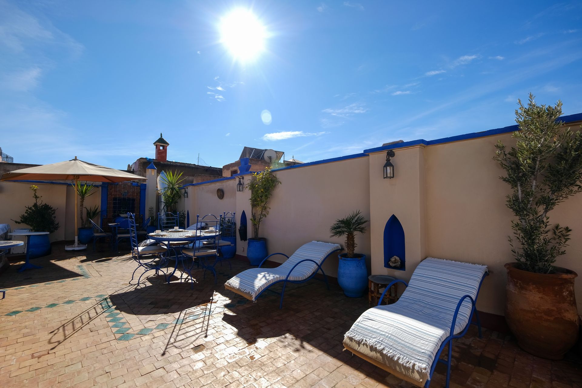 Rooftop terrace with lounge chairs, dining table, and blue accents, under a bright, sunny sky.