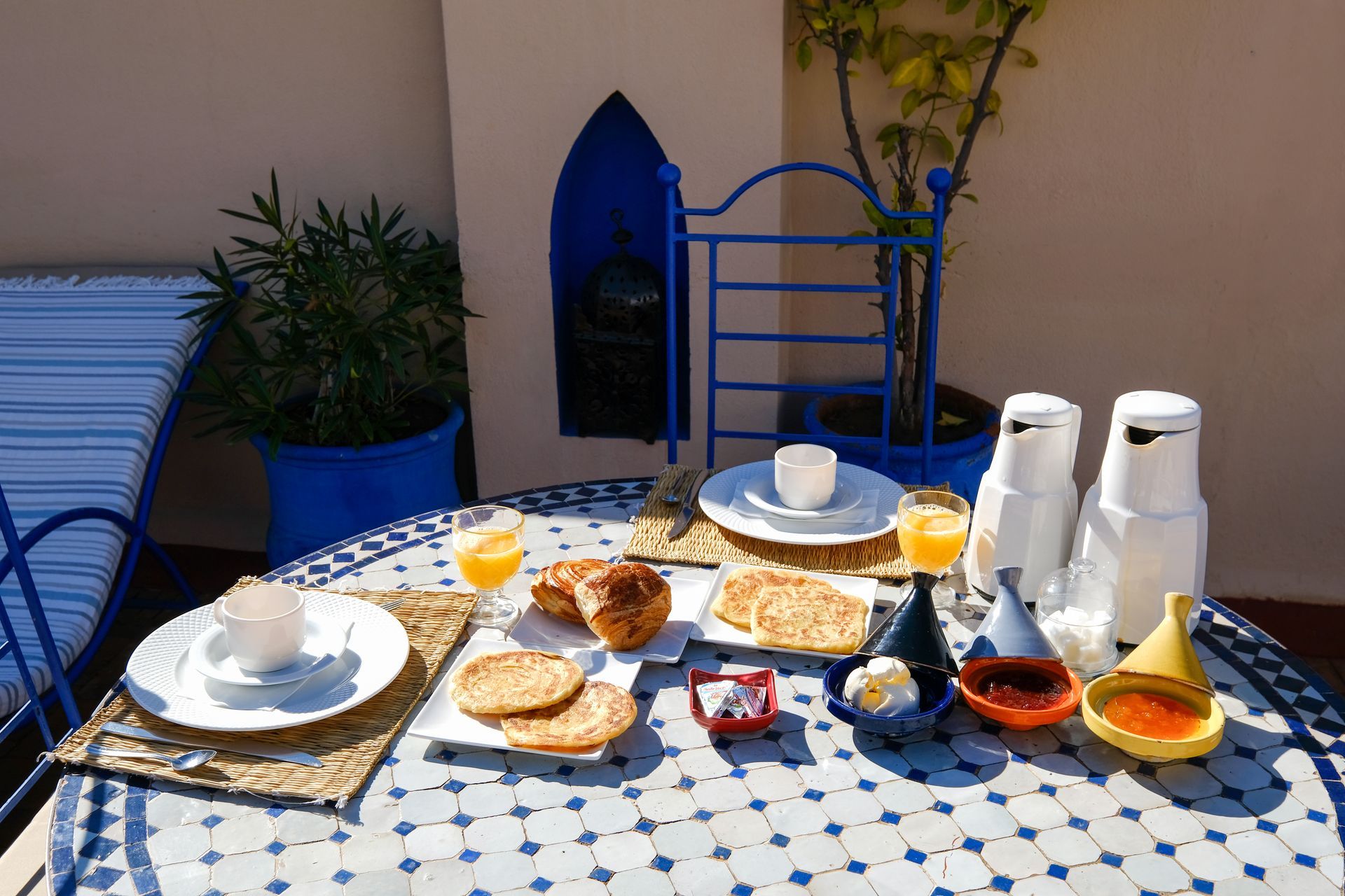 Breakfast on a blue tiled table: pastries, juice, and condiments, on a sunny patio.