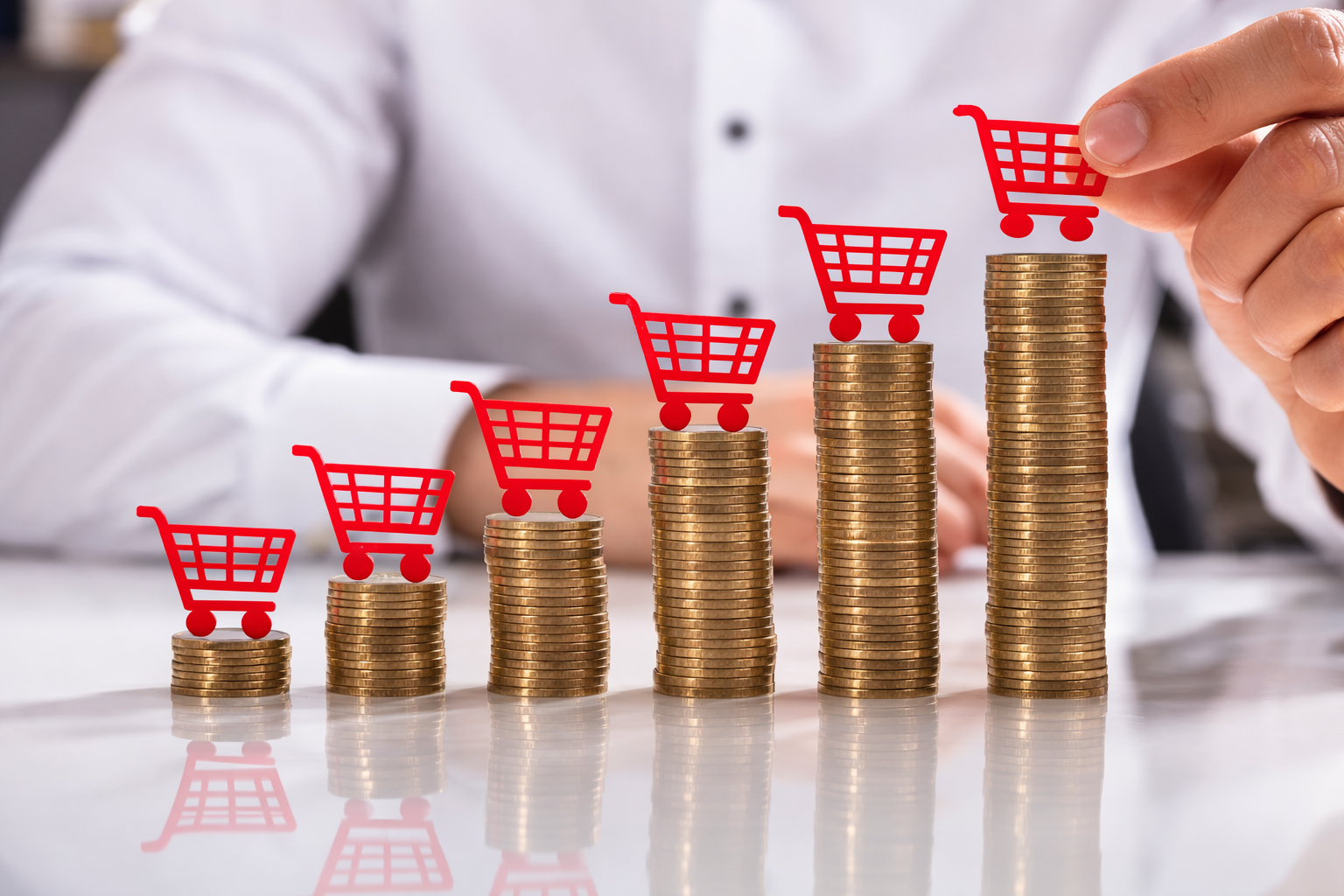Uma pessoa colocando um ícone de carrinho de compras vermelho em uma pilha crescente de moedas de ouro, representando o aumento dos custos para o consumidor.
