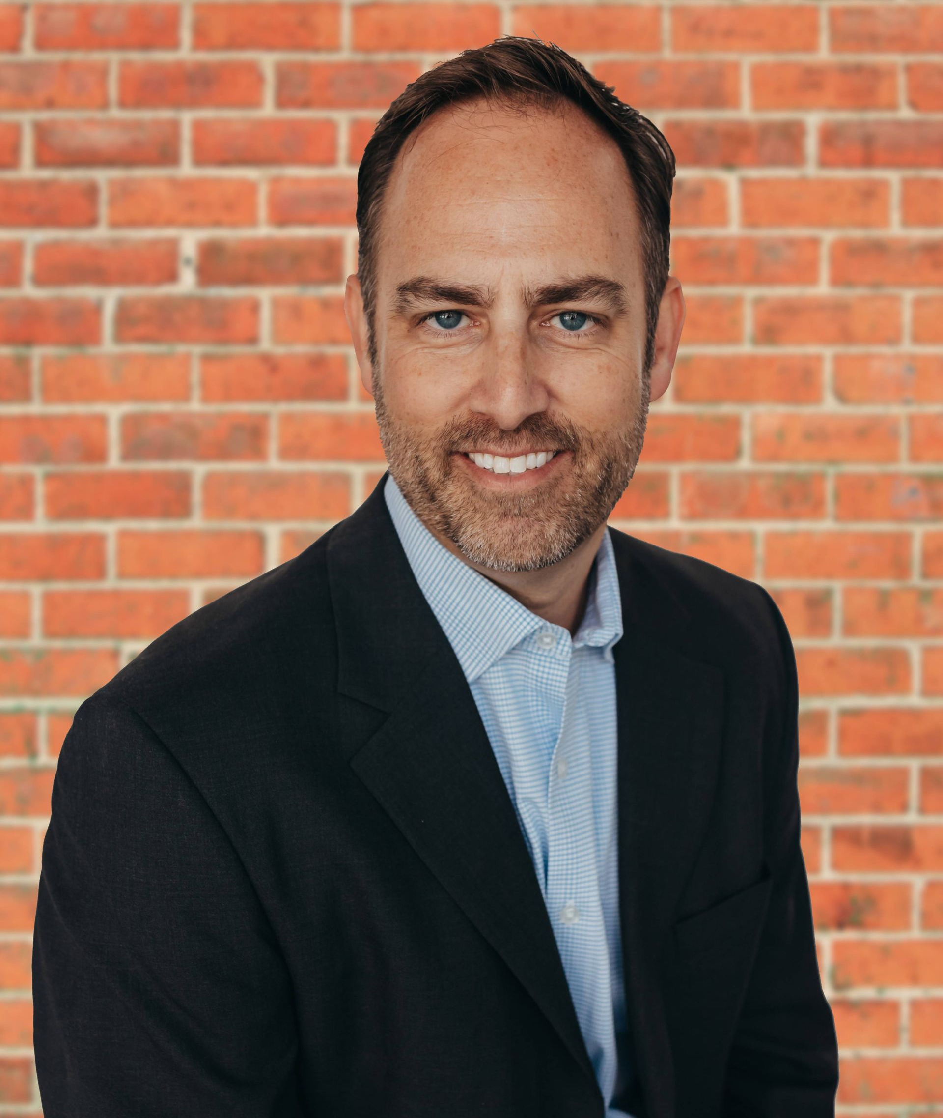 Man in suit smiling, light blue shirt, neutral background.