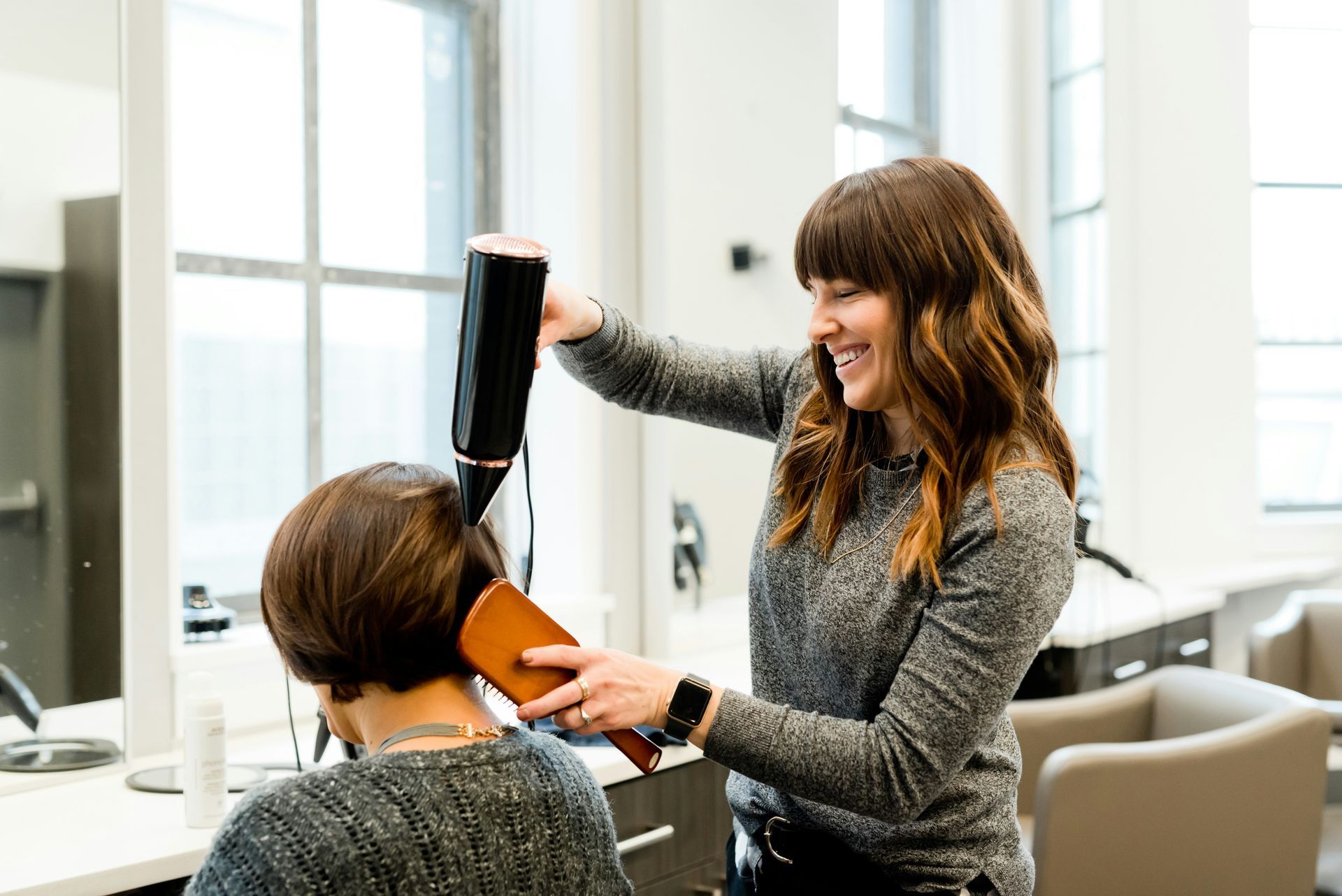 Hairdresser blow-drying a client's hair in a salon. Smiling woman with dark hair, using a hair dryer and wooden brush.