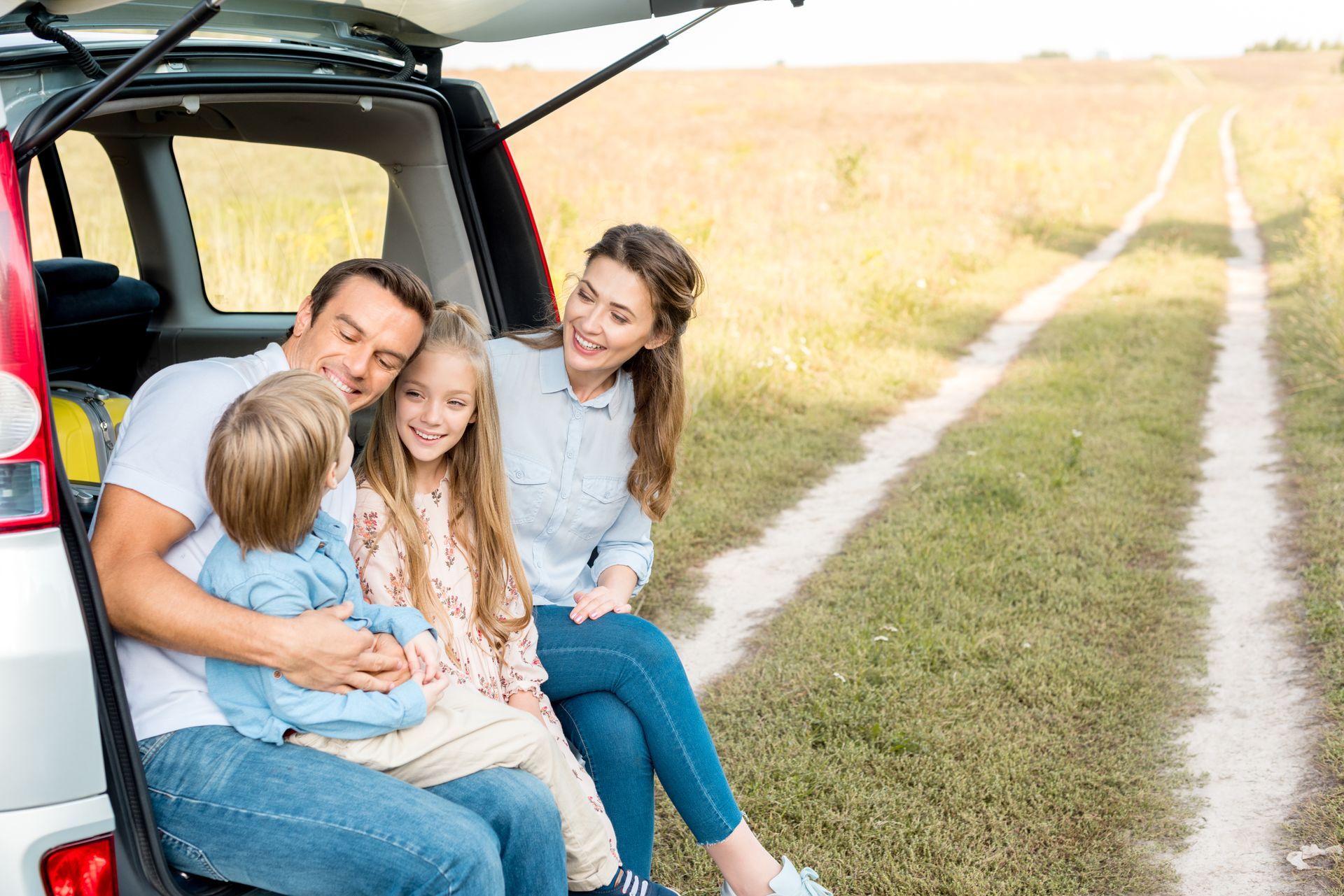 Family sitting in a car trunk outdoors, smiling. Two children are between their parents, a dirt road stretches ahead.