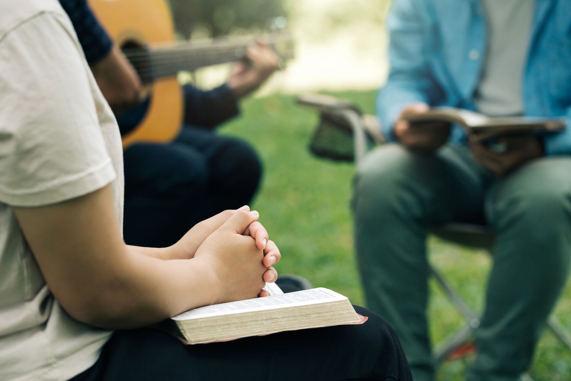 Person with clasped hands resting on a Bible, with friends; one plays guitar, another reads, outdoors.