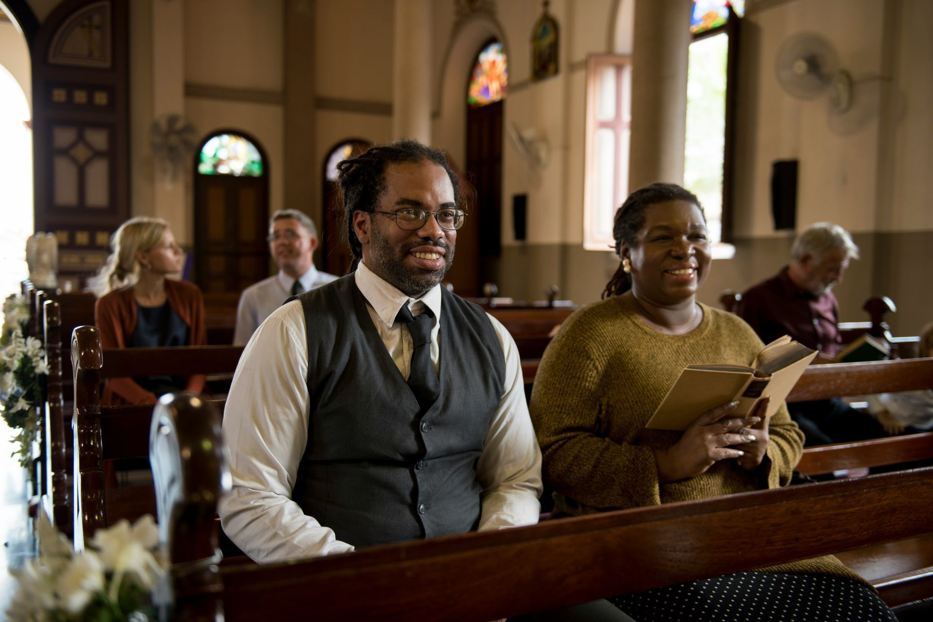 A Black man and woman smile while sitting in church pews, holding prayer books. Others sit nearby.