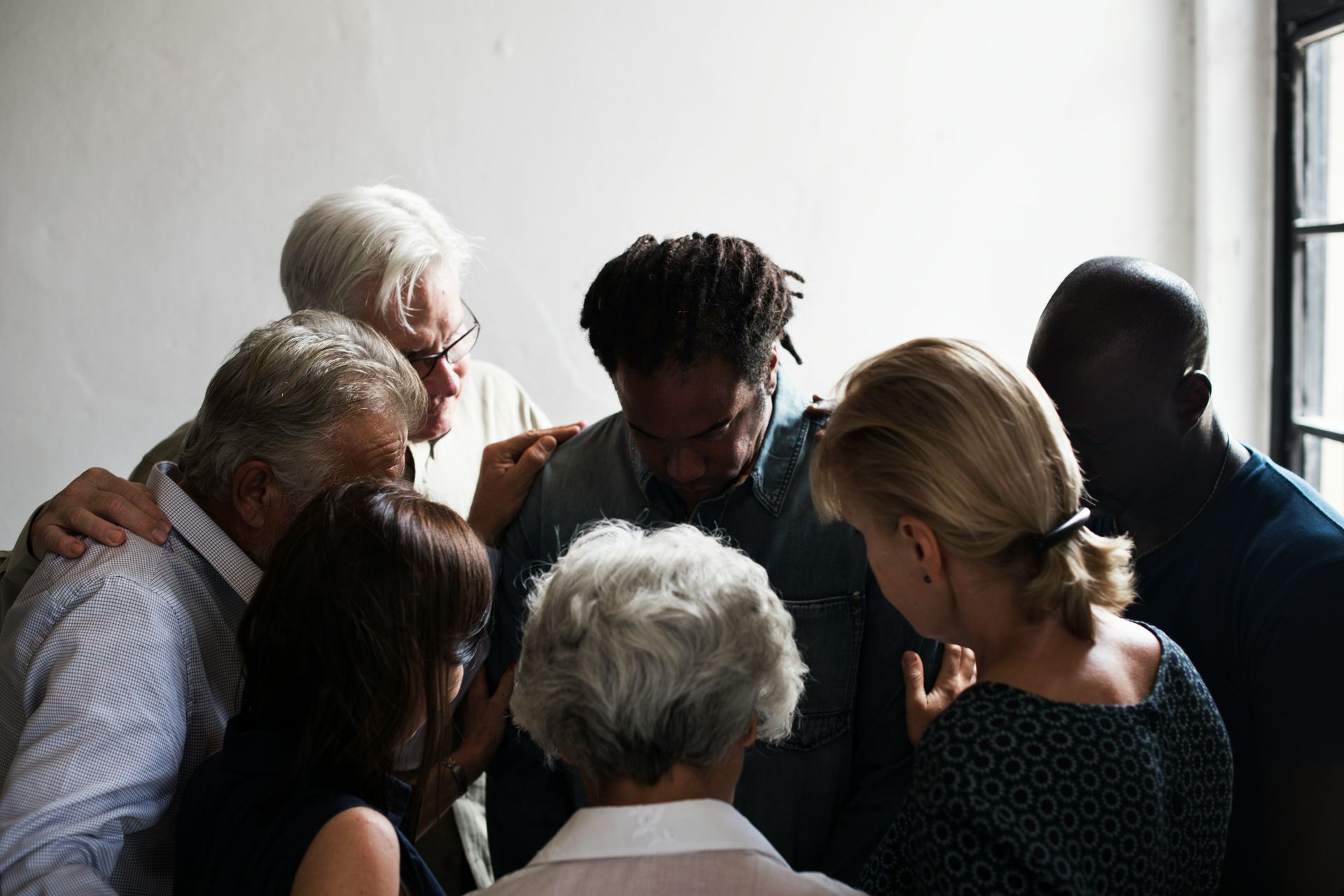 Group of diverse people praying together, with hands on each other's shoulders, in a room with a window.