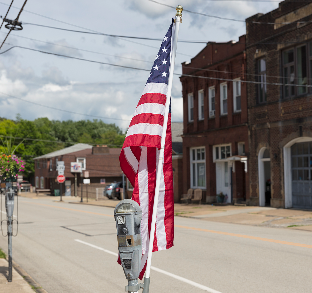 American flag on a pole stands on a street in a small town, with brick buildings in the background.