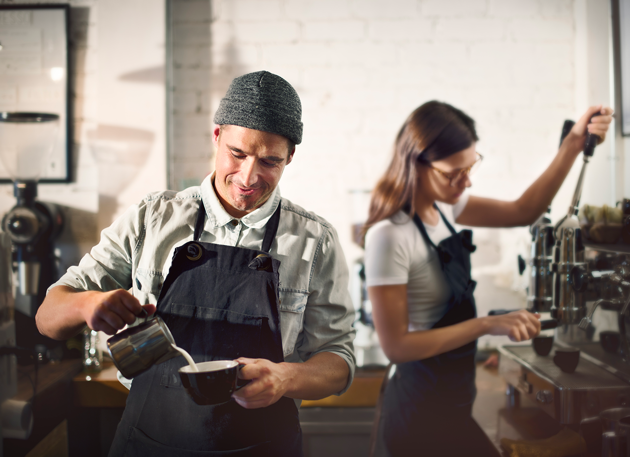 Two baristas making coffee, one pouring milk, the other operating the espresso machine.