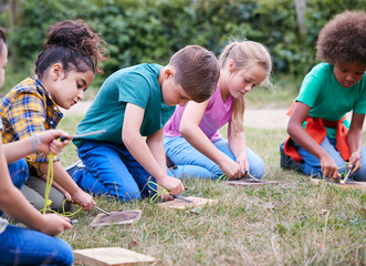 Children kneeling outdoors, carving wood with small tools on wooden blocks.