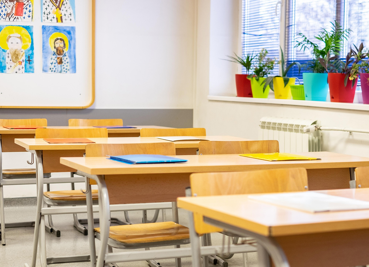Empty classroom with desks and chairs, colorful plants on a windowsill, and artwork on the wall.