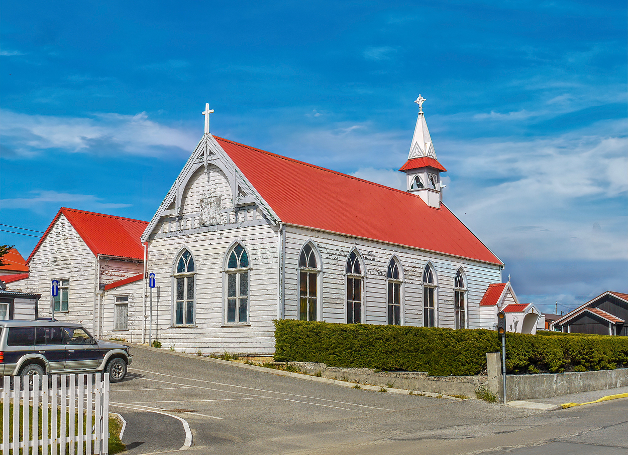 White church with red roof, blue sky, parked vehicle, and hedge.