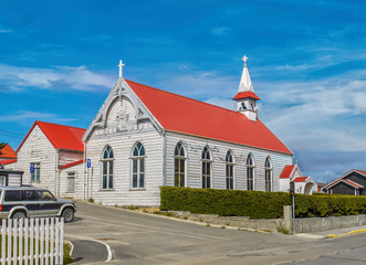 White church with red roof, blue sky, parked vehicle, and hedge.