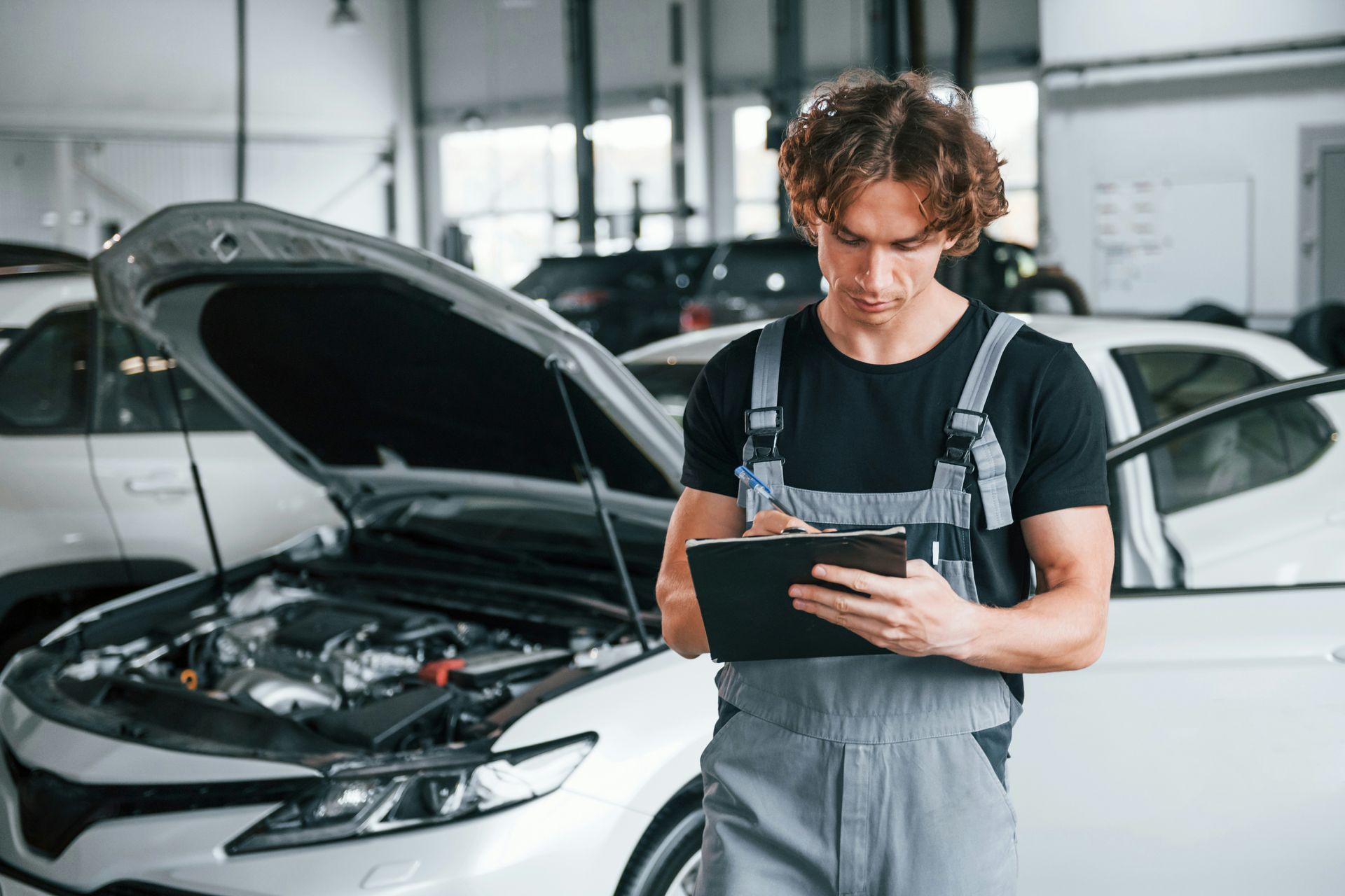 Mechanic in gray overalls and black shirt inspecting a car with open hood, writing on a notepad.