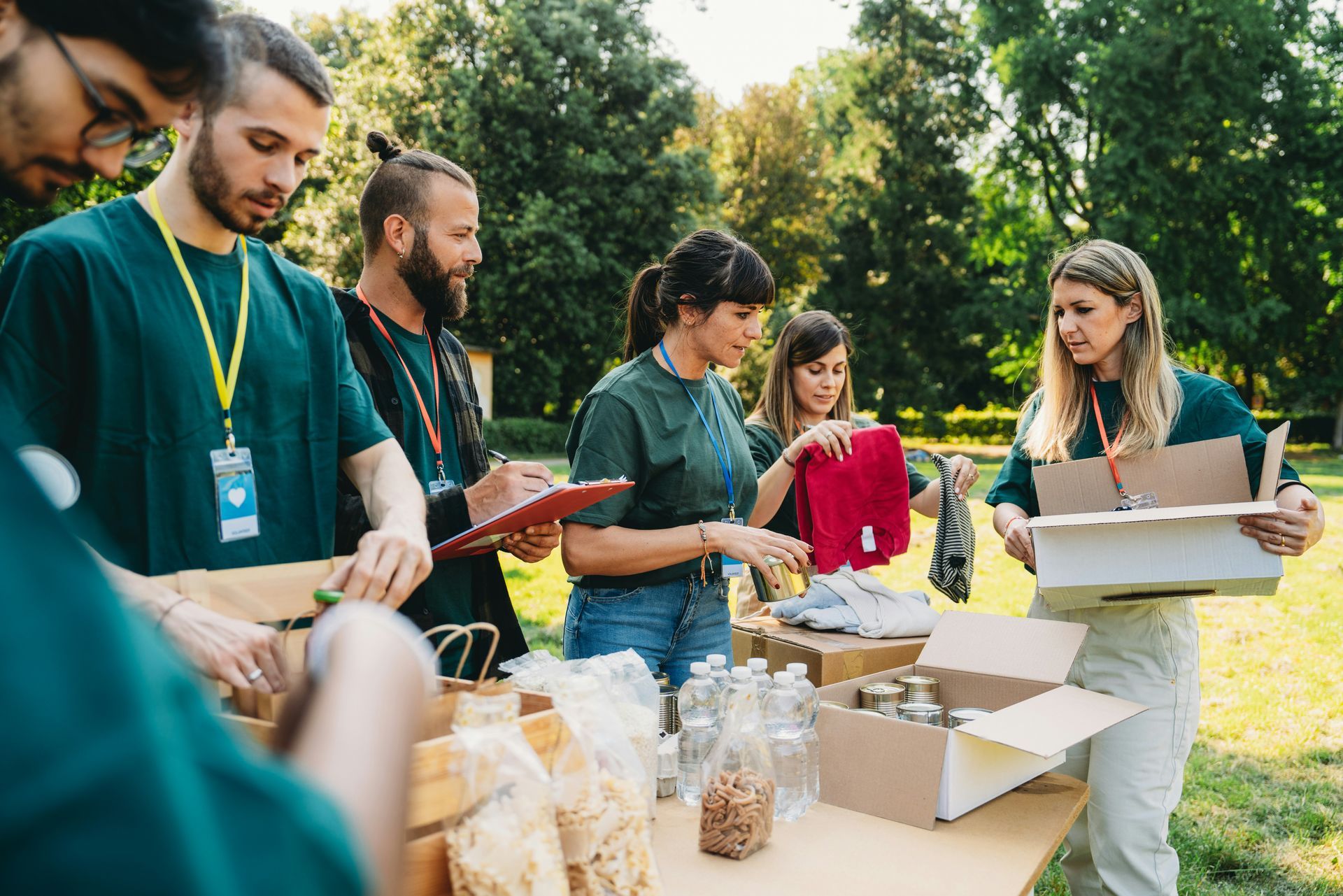 Volunteers packing food donations outdoors at a table in a park, wearing green shirts.