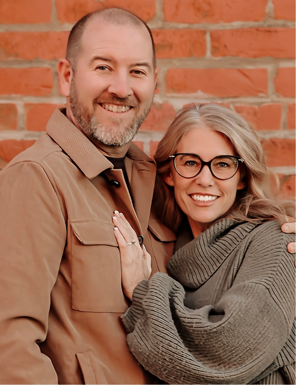 Man and woman smiling, posing by a red brick wall. The man wears a tan jacket; the woman, a gray sweater.