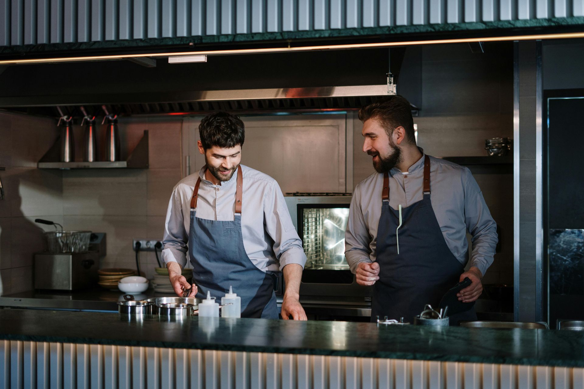 Two chefs wearing aprons work behind a bar. One looks at the other, smiling.