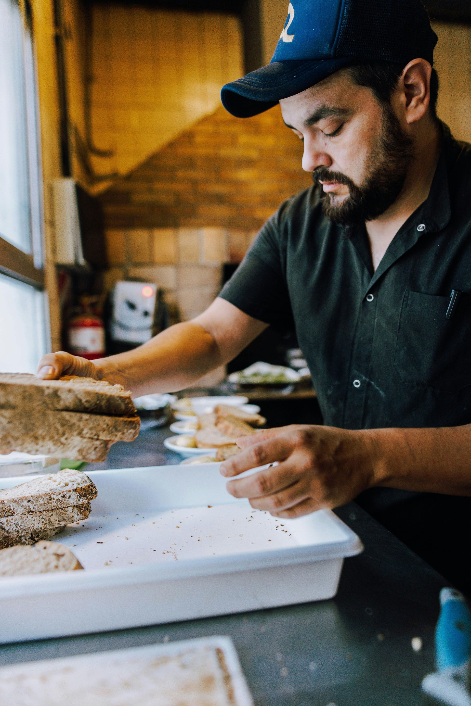 Chef in blue cap, preparing food in a kitchen, leaning over a white tray with bread.