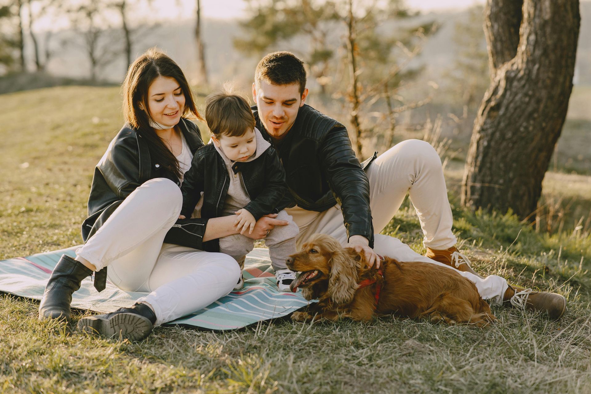 Family of three and dog on a blanket in a park, smiling.