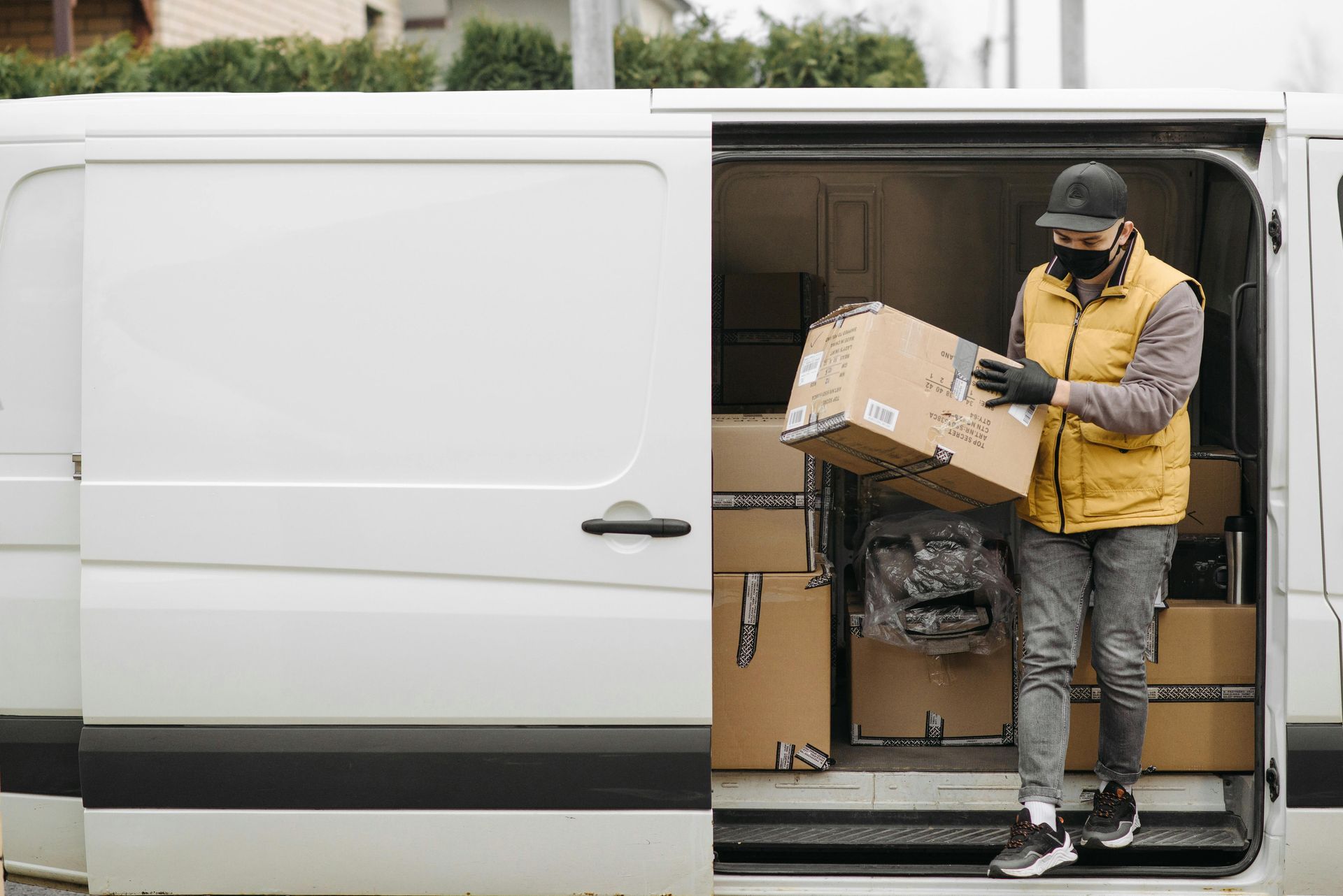 Delivery person in mask, gloves, and yellow vest, unloading cardboard box from white van.