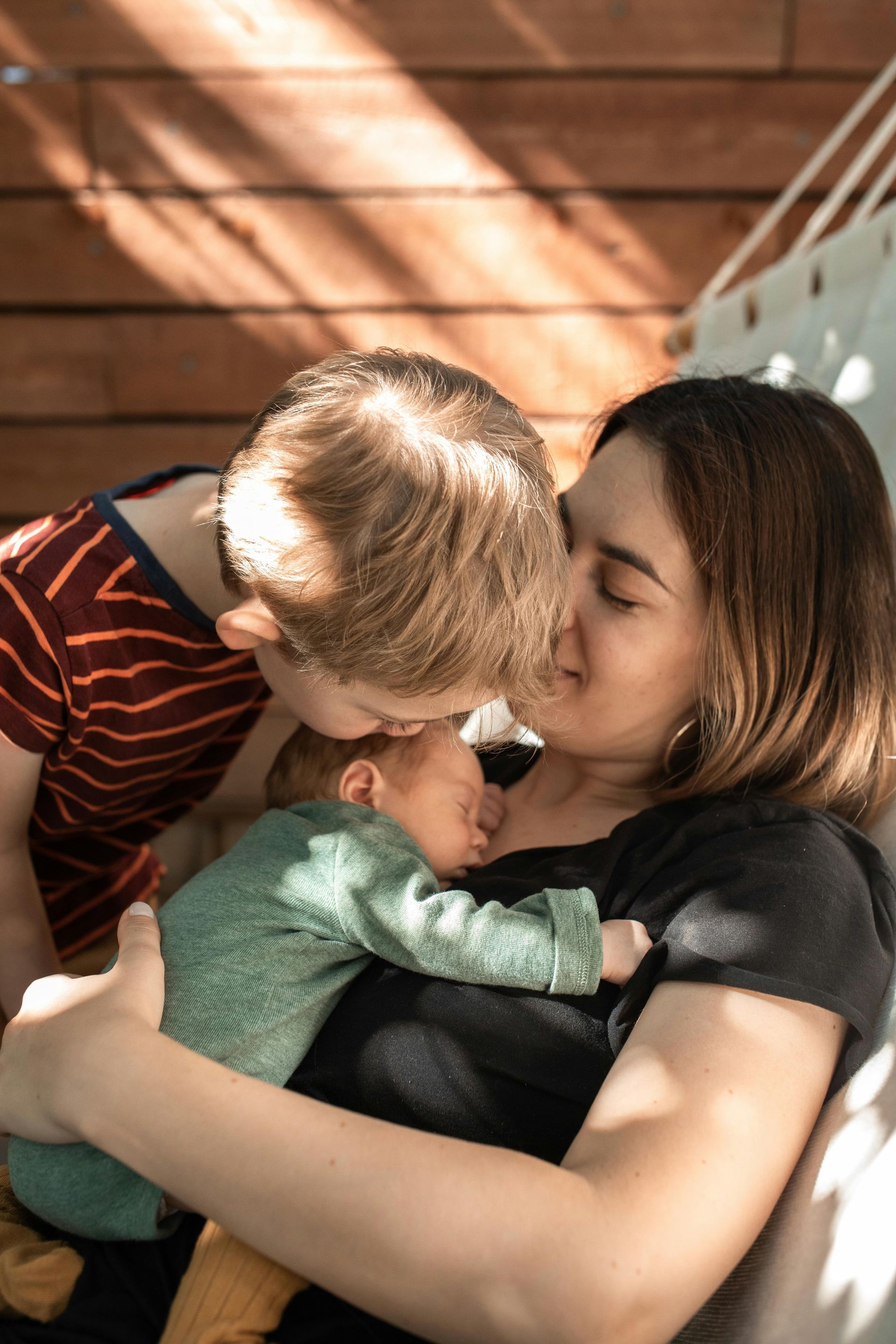 A mother cradles a newborn as a person leans in to kiss the baby, who sleeps, outside.