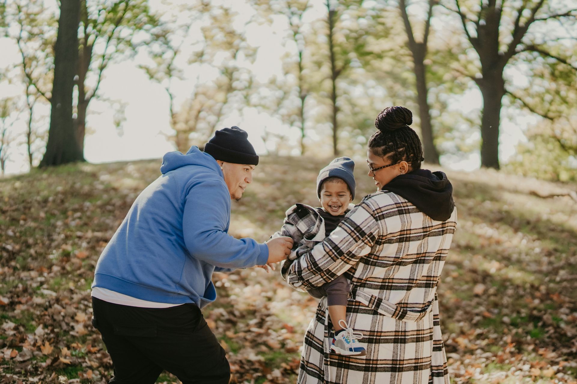Family in a park. Man in blue jacket, woman in plaid coat holding a toddler. Autumn leaves.