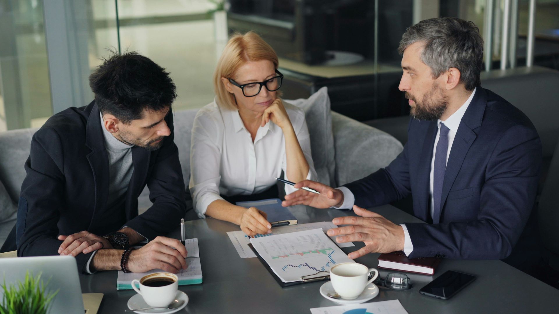 Three business people review data at a table. One gestures, two look on; coffee cups and laptop present.